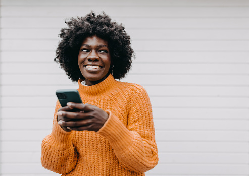 A cheerful African-American woman in her early twenties, with a stylish afro hairstyle, stands against a white background. Engrossed in her mobile phone, she's using a virtual assistant app with excitement, demonstrating the perfect blend of technology and beauty.