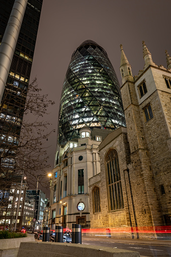London. UK-02.02.2024. A night time street view of The Gherkin building in the City of London with St. Andrew Undershaft Church in front.