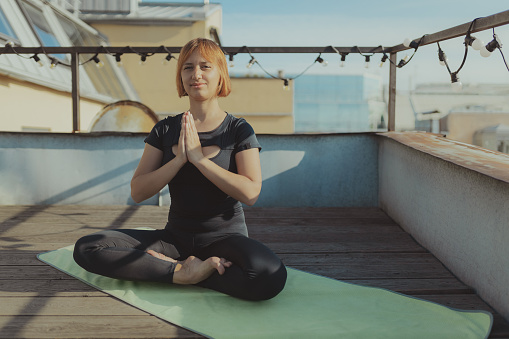 Portrait of young girl in black sportswear sits in position of yoga meditates on green carpet on terrace roof, concept yoga and meditation in urbanistic city