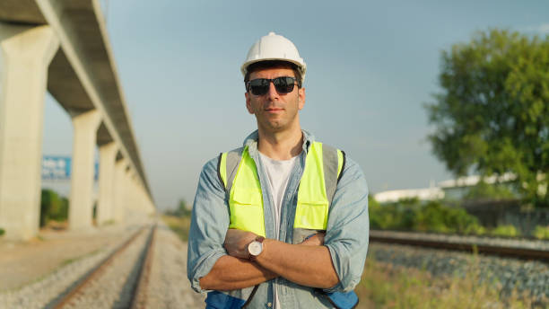 istock Handsome engineer man or architect in safety uniform and helmet standing with arms crossed on railroad smiling and looking at camera. Engineer leadership concept 1992362464