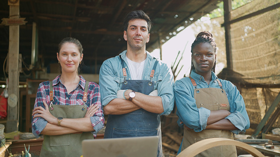 Portrait of three confident carpenters man and woman standing with arms crossed looking at camera. Team of professional carpenters in workshop. Carpenter and woodcraft concept