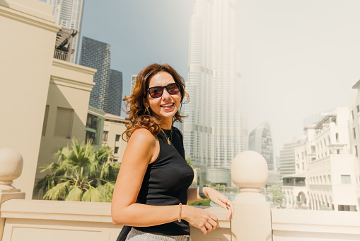 Portrait of smiling west asian woman with shopping bag walking the central district of Dubai enjoying view of Skyscrapers by the waterfront during sunny day