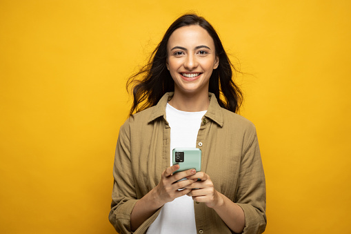 Portrait of cheerful positive modern woman using phone isolated yellow background