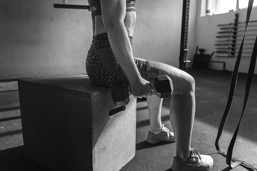One woman athlete sits on a gym box, performing strength exercises with dumbbells