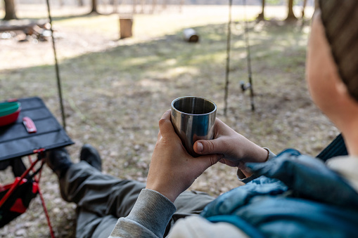 Close-up of a metallic mug in hands of a man sitting at the foldable touristic table at the campsite in the woods