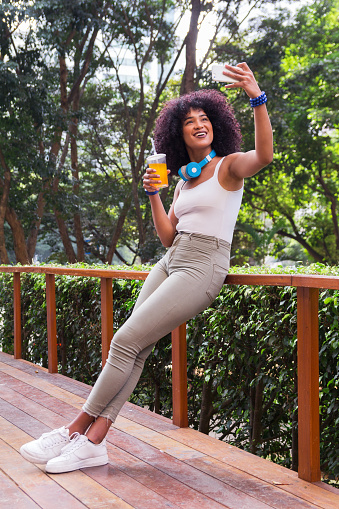 Full length body of beautiful black girl taking a selfie photograph and cup of juice for social media. Woman is smiling and leaning against railing. Wooded background.