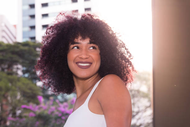 Portrait of woman looking to the side and smiling. Builindgs and tree tops. She is black, on her early twenties, frizzy hair.