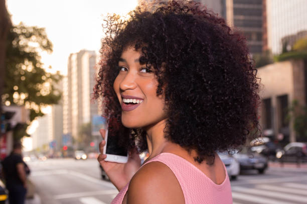 Sideways of woman with smartphone in the hand in the city during summertime. She is black, on her early twenties, Afro style frizzy hair.