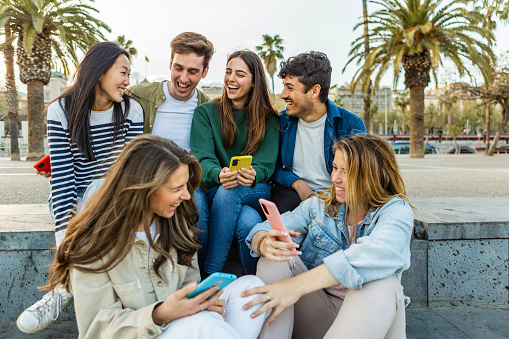 Happy group of student people with mobile phones having fun sitting outdoors. Millennial generation diverse friends laughing enjoying time together social gathering in city street. Youth lifestyle.