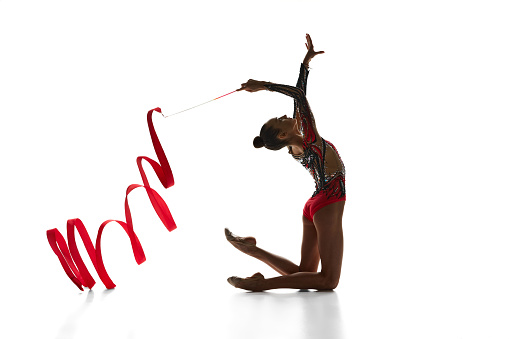 Artistic, talented teen girl, rhythmic gymnast performing with red ribbon, dancing against white studio background. Concept of sport, beauty and grace, competition, art, youth, hobby
