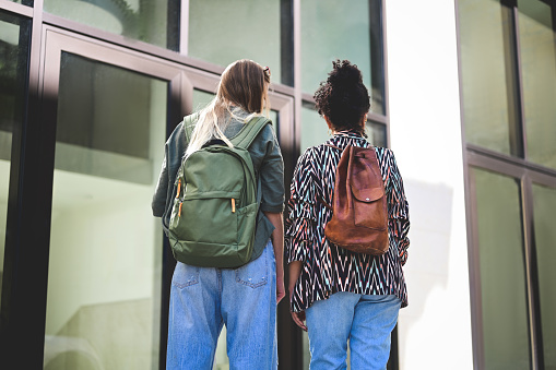 Rear view of two female friends with backpack in front of a campus.