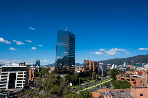 The photograph is a panoramic portrayal of Bogotá, captured at midday under a clear, azure sky. It presents a vivid tableau of the city's modern architectural landscape, where contemporary buildings bathed in bright sunlight stand in stark contrast against the backdrop of the surrounding mountains. These modern structures, with their sleek designs and reflective surfaces, glimmer under the midday sun, showcasing the city's urban development and architectural advancements. The sky, a brilliant shade of blue, stretches overhead, providing a serene and expansive backdrop to the bustling cityscape below. The surrounding mountains, a defining feature of Bogotá's geography, encircle the city, adding a sense of grandeur and natural beauty to the urban setting. The photograph captures the dynamic interplay between nature and urbanization in Bogotá, highlighting the city's unique position where modernity meets the majesty of the natural world.