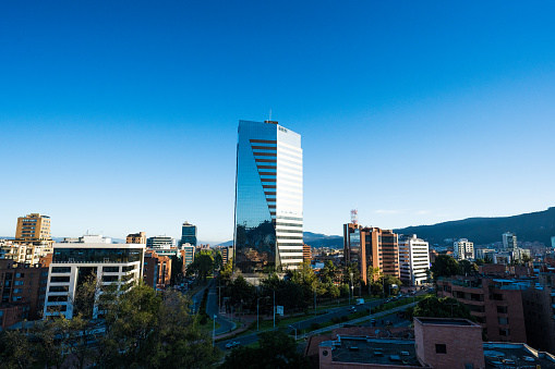 The photograph presents an exquisite juxtaposition of modernity and nature in Bogotá, Colombia. Captured in the early hours of the morning, it features a striking, contemporary glass building that glistens under the emerging sunlight. The building's sleek, reflective surfaces mirror the vibrant blue of the morning sky, creating a mesmerizing visual harmony. In the backdrop, the majestic Andean mountains stand tall, draped in a serene mist, providing a contrasting backdrop of natural ruggedness to the building's polished urbanity. The sun, just peeking over the horizon, bathes the scene in a soft, golden light, highlighting the building's geometric contours and the mountains' imposing presence. This photograph eloquently encapsulates the blend of modern architecture and timeless natural beauty that characterizes Bogotá.