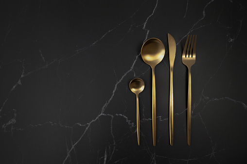 Top view of gold cutlery on black marble background. Fork, knife, spoon and dessert spoon. Table setting, flat lay, copy space.