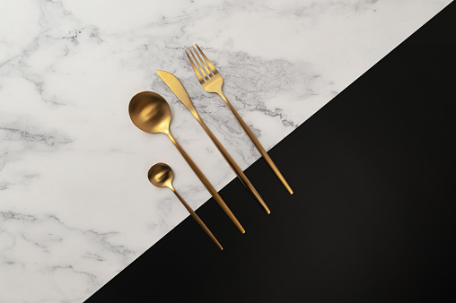 Top view of gold cutlery on white marble and black background. Creative table setting flat lay. Fork, knife, spoon and dessert spoon. Copy space.