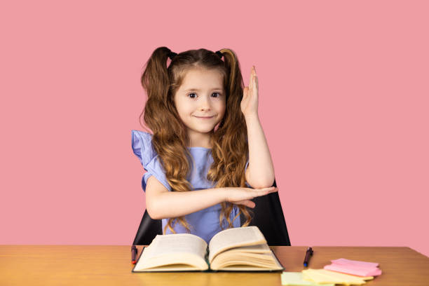First day at school. A happy smiling schoolgirl raised her hand while sitting at a table with the book in front of her on a pink background. High quality photo