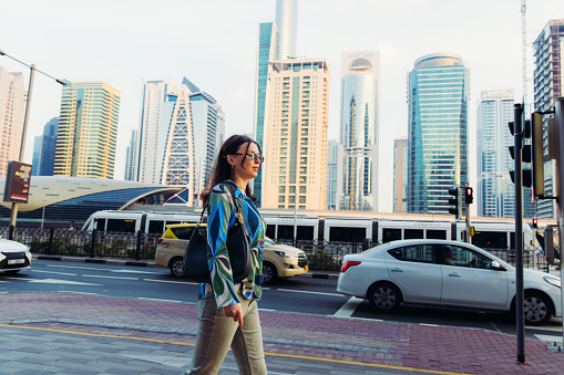 Handsome businesswoman in sunglasses with handbag walking from a job on the street during rush hour with a view of Dubai city skyline