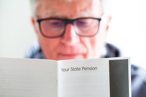 A retired senior man in his 70s reads information about receiving his pension. Focus on the leaflet with the man slightly defocused in the background.