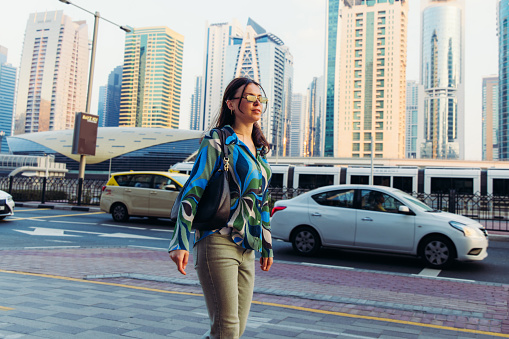 Handsome businesswoman in sunglasses with handbag walking from a job on the street during rush hour with a view of Dubai city skyline