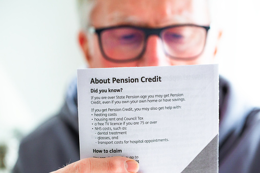 A retired senior man in his 70s reads information about pension credit. Focus on the leaflet with the man slightly defocused in the background.