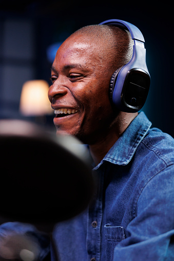 Closeup of joyful african american man wearing headphones and speaking via a microphone moderates online exchanges. Cheerful black guy using podcast equipment to broadcast a live radio show.