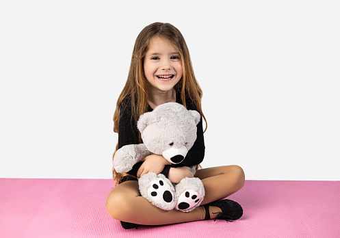 Smiling little girl dressed in black gym clothes, holding a gray teddy bear in her arms, sitting on a pink yoga mat, isolated on a white studio background. High quality photo