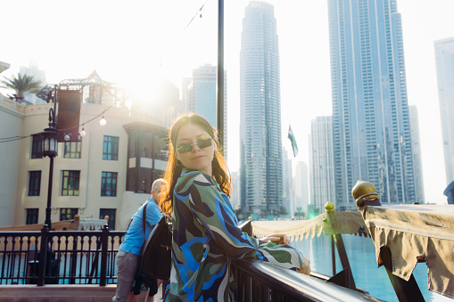 Side view of beautiful businesswoman in sunglasses holding handbag contemplating soft sunshine over modern buildings and the bay in the central business district of Dubai, United Arab Emirates