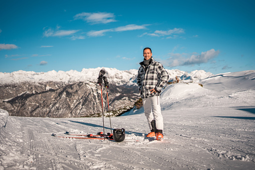 Mid adult Asian male skier standing on a snowy mountain slope with ski equipment. He is wearing casual winter clothing and a helmet, with a scenic mountain range in the background.