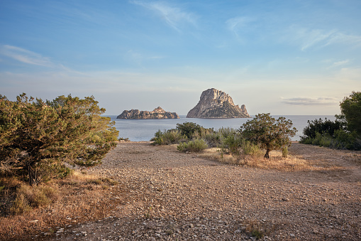 Wide-angle view of a flat gravel ground overlooking Cala d'Hort, a bay of water located on the southern coast of Ibiza, renowned for its crystal-clear waters and its scenic view on the iconic rocky isles of Es Vedrà and Es Vedranell. The delicate warm light of a Mediterranean summer sunset, picturesque clouds, deep blue waters as far as the eye can see, a wide variety of lush Mediterranean shrubs and plants — juniper, rosemary and thyme among the others. High level of detail, natural rendition, realistic feel. Developed from RAW.