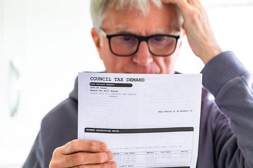 Portrait of a senior man in his 70s checking his council tax bill at home in his office. He has a worried expression and touches his face with his hand while looking at the bill. Focus on the tax bill with the man's face defocused beyond.