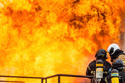 Firefighters protect themselves with water from their hoses against a large flame resulting from an explosion