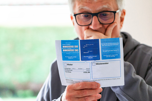 Portrait of a senior man in his 70s checking his water bill at home. He has a worried expression and touches his face with his hand while looking at the bill. Focus on the bill with the man's face defocused beyond.