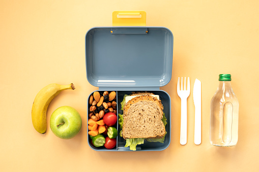 School lunch box with sandwich, vegetables, fruit and water on yellow background, healthy eating concept, top view