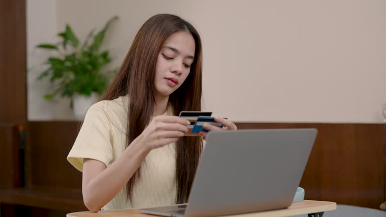 Asian lady with laptop, comparing credit cards, online shopping, decision-making at home. Thoughtful woman analyzing credit cards, engaging in e-commerce, serious financial consideration