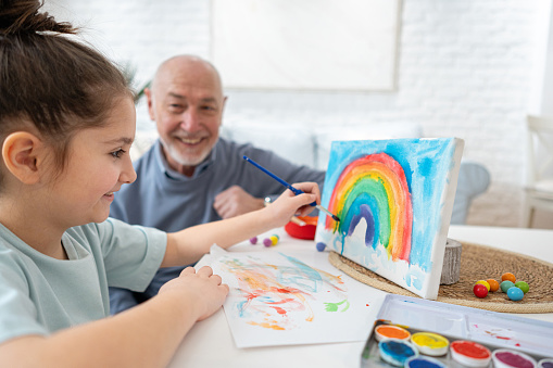 Young Caucasian girl, painting on canvas with watercolor paint, while having an great playtime with her grandfather