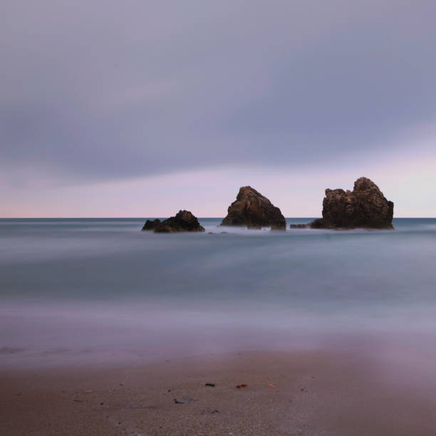 captured with long exposure technique, a rock in the middle of the sea surrounded by a pink-toned sky and a blue sea. stock photo - long exposure rock cloud sky photos et images de collection