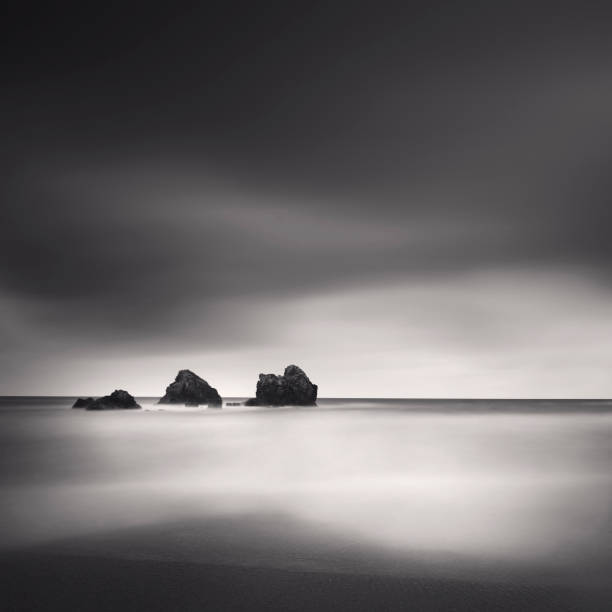 a black and white photograph taken with long exposure technique featuring three small rocks, a smooth sea, and a dark sky. stock photo - zen like sea horizon over water blurred motion ストックフォトと画像