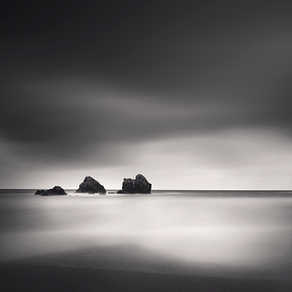 A black and white photograph taken with long exposure technique featuring three small rocks, a smooth sea, a dark sky, minimalist style, and a serene atmosphere.