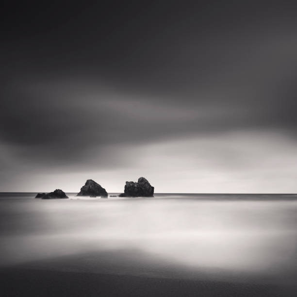 a black and white photograph taken with long exposure technique featuring three small rocks, a smooth sea, and a dark sky. stock photo - black and white landscape square long exposure - fotografias e filmes do acervo