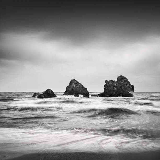 a black and white photograph taken with long exposure technique featuring three small rocks, a smooth sea, and a dark sky. stock photo - zen like sea horizon over water blurred motion ストックフォトと画像