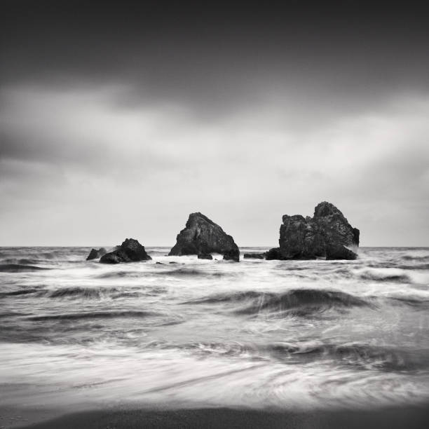 a black and white photograph taken with long exposure technique featuring three small rocks, a smooth sea, and a dark sky. stock photo - black and white landscape square long exposure стоковые фото и изображения