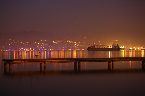 At night, the reflections of city lights shimmering on the sea, with a large tanker docked behind the pier. stock photo