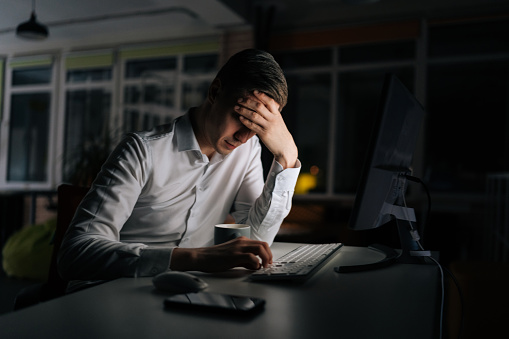 Exhausted businessman working in evening office alone. Tired anxious manager losing failure working on computer at late night. Tired worried business man at workplace in office holding head on hands.
