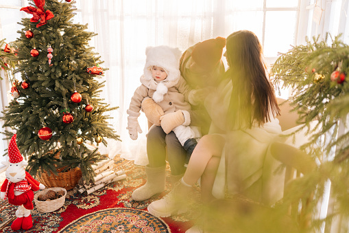 Remote view of happy family wearing winter clothes sitting in decorated room of summer house on Christmas eve. Young mother and father with loving child baby having fun. Family gathering together.