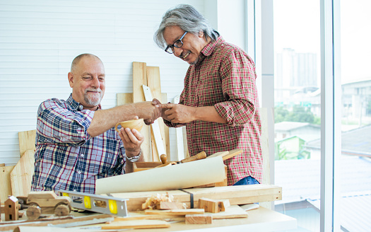 Two senior mixed race diverse happy retired old male woodworkers or carpenters wearing check shirts, creating DIY wooden car toy together for decoration and furniture. Hobby, Retirement Concept