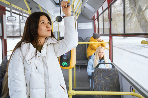 Portrait of cute nice female with long dark hair wearing white coat dreaming and thinking about future plans. Other passengers sitting staring at phone and watching city life through tram windows.