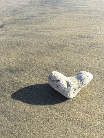 On a smooth brown and black highlighted sandy beach sits a solitary white stone heart casting a shadow to the left.