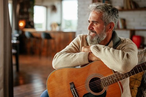 A thoughtful mature Caucasian man is at home with his acoustic guitar and looking through the window.