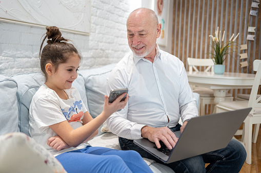 Senior Caucasian man using laptop, while his granddaughter using mobile phone