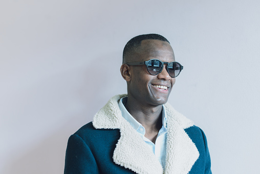 A male black model stands wearing sunglasses and a blue jacket, looking towards the camera over light solid background high res image